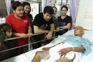 ภาพ นายวิชัย ตราชื่นต้อง  บนเตียงนอนคนไข้ในโรงพยาบาล มีคนเยี่ยมยืนอยู่ข้างเตียง 6 คน